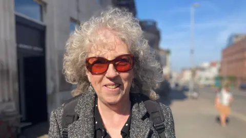 Claire Hamilton/BBC Sue smiles at the camera. She has grey curly hair and wears sunglasses, a grey jacket and a black and white polka dot blouse