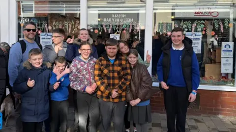 BBC The photo shows a group of people smiling at the camera. They are all standing in front of a shop, which reads 'St. Anne's Charity Shop' in bold black letters. 
