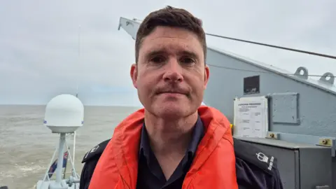 BBC / Natalie Glanvill Andy Vidamour is standing on a grey boat. He has short brown hair and is wearing a navy blue shirt with crown logos on lapels. He is wearing an orange life jacket.