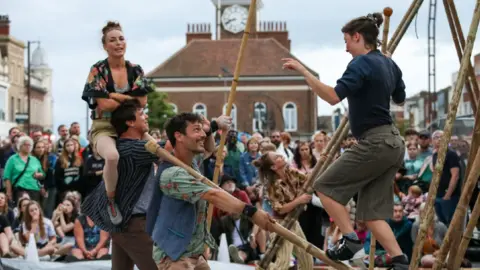 Stockton Borough Council Performers balance on wooden poles in front of a crowd.