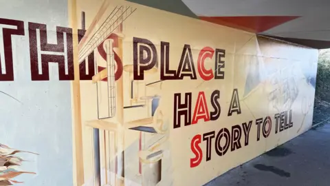 BBC/RICHARD EDWARDS This is a picture of a mural, painted at Castleford's Tickle Cock Bridge, which is one of the main routes into the town centre. In big, bold letters it says 'This Place Has a Story to Tell.' Cream, red and brown colours have been used. An outstretched palm is just visible to the right of the image.