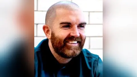 Chris Chris with dark brown shaved hair and a brown beard wearing a dark blue shirt. There is a pale brick wall in the background. He is looking to the side of the lens and is laughing.
