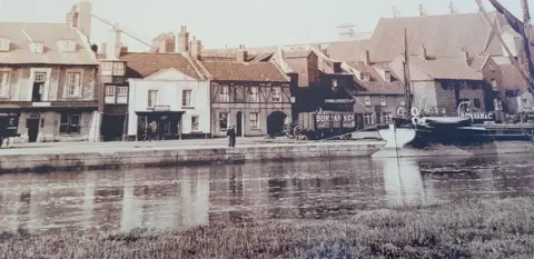 Marcus French Image of Wells Harbour from around the time that French's fish and chip shop moved on to the waterfront. In the foreground is the water and then a line of shops and buildings along the quay. A boat is in the right hand side of the frame. 