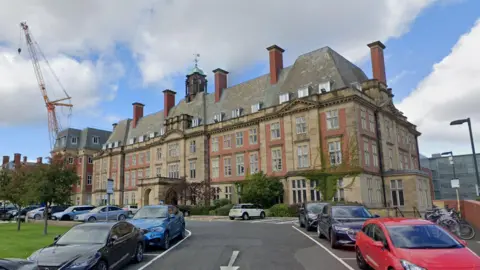 An external view of Peacock Hall which is part of the Royal Victoria Infirmary (RVI). The building is made of red brick in a similar style to grand French houses and has ivy growing up the walls. Cars are parked outside. Six of the building's chimneys can be seen. In the middle of the roof stands the lantern toped with a copper dome and weather vane.