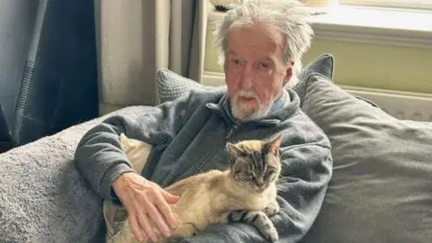 William Brindley, 77, wears a grey zip-up fleece and strokes a cat resting on his shoulder. William has grey hair which sticks out and he has grey bread. He is sitting on a grey sofa. 