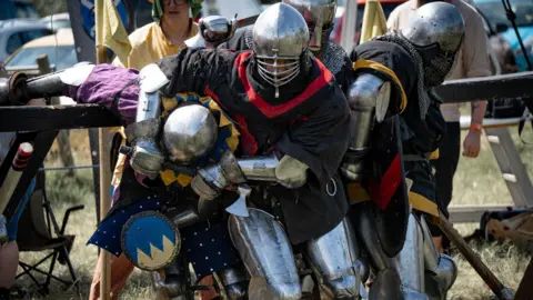 David Polier Two people in suits of armour engaged in combat. One has the other in a headlock and people dressed in medieval clothing are looking on