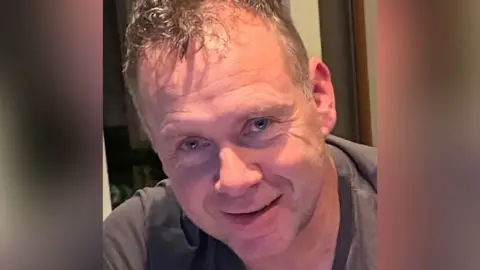 A man with thinning grey hair and a grey t-shirt smiling at the camera - only his head and shoulders are pictured.