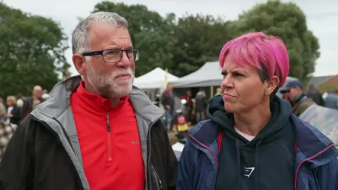 A man with short grey hair is wearing a black coat over a red top with a three-quarter zip. A woman with short pink hair is standing next to him and is wearing a blue hoodie underneath a blue coat 