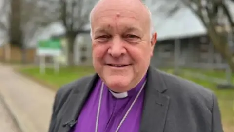 A picture of David Williams smiling at the camera. He is wearing a purple shirt with a grey jacket. 