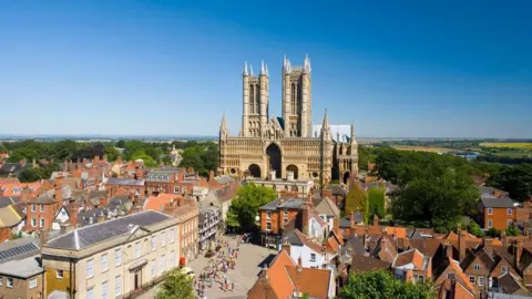 An aerial view looking out across the castle square in Lincoln from the castle to Lincoln Cathedral. Several buildings are dotted around the castle square and Lincoln Cathedral with its two towers stand looking out at the city. The sky is blue and greenery and fields can be seen in the distance.