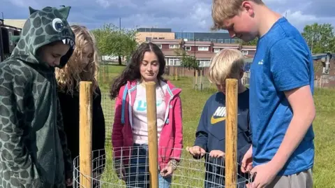Five youngsters looking at the spot where they had planted a fruit tree. The planting spot has metal mesh and three wooden poles. It is on a field with other similar planting structures dotted around in the distance. There are a number of relatively modern two-storey terraced houses on the edge of the green.