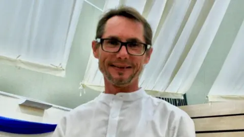 Emanuel with brown hair wearing a white shirt and black glasses in an office. He is smiling.