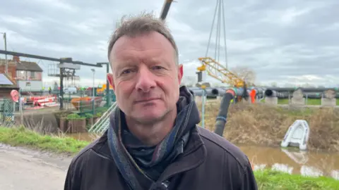 Ian Withers stands in front of large pipes and pumps being installed along a river on the Somerset Levels. He is in his 40's and wears a scarf over a black fleece. There are men working behind him.