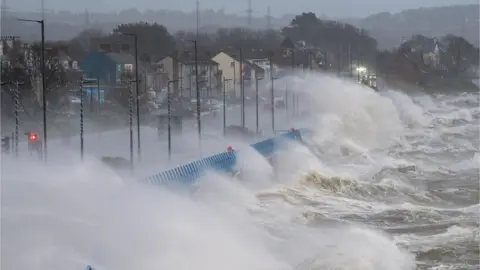 A stormy sea hitting a blue gate. The sky is grey and the spray from the sea is going over the blue gates toward a row of buildings. 