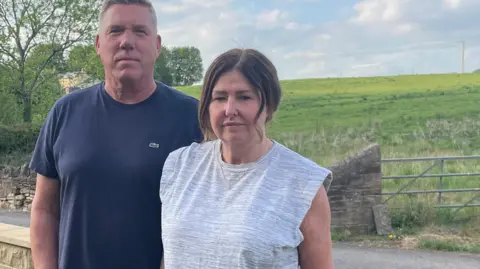 Tom MacDougall/BBC A man in a navy blue t-shirt stands next to a woman with dark hair wearing a grey t-shirt. Behind them is a wall and a gate, with an open field beyond.
