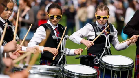 Getty Images Close-up of a group of teenage girls who are marching drummers performing outdoors. They are wearing traditional outfits and their drumsticks are raised, about to come down on the drums. Spectators are blurred in the background