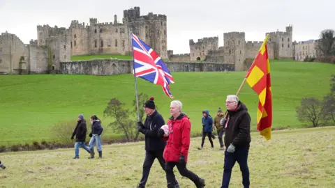 PA Media Three men are walking on the fields in front of Alnwick Castle. Two are holding large flags and the one in the middle is carrying a football.