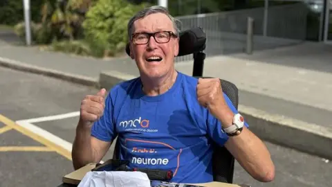 A man in a blue shirt sitting in a wheel chair. He is looking at the camera and lifting up his arms. He is wearing glasses.