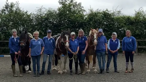 A row of eight volunteers in blue polo tops stand in a row at Watershed RDA with three ponies in between them