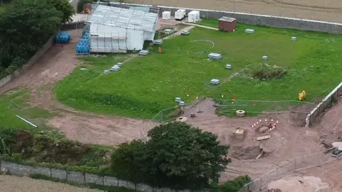 A field surrounded by a wall with a building of scaffolding inside and the field dotted with uknown objects and wear from tyres.