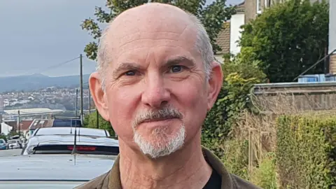 LDRS A man is standing outdoors on a residential street with houses, parked cars and trees in the background.