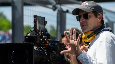 PA Media A man in a blue shirt, black cap and sunglasses holds up his hand behind a large black camera.
