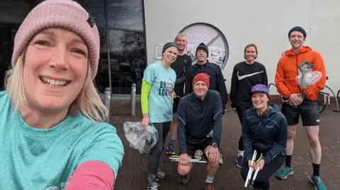 A selfie with a close-up of Claire, smiling to camera, and seven other ploggers dressed for running and holding plastic bags and litter sticks