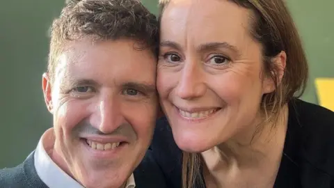 Aurelie Boas A man with brown hair and moustache and a woman with brown hair. They are smiling. Behind them is a green wall. 