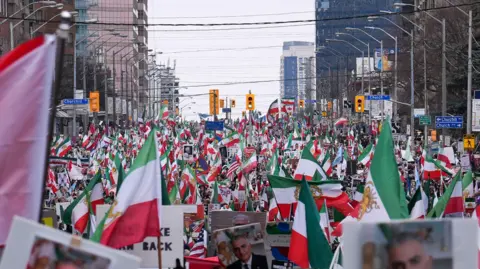 Andolu via Getty Images A sea of people, carrying Iranian flags at at Yonge Street in Toronto, Canada. The sky behind the protesters is grey.