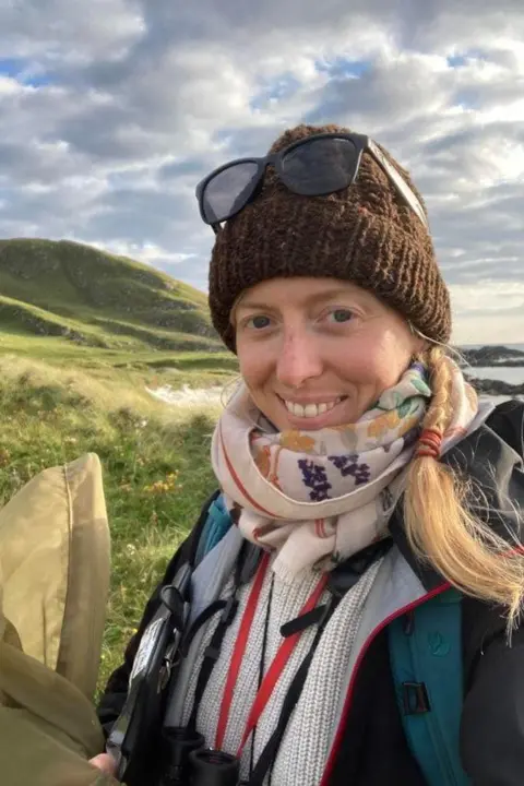 University of Aberdeen A blonde haired woman is wearing a brown woolen hat with sunglasses perched on top. She is smiling and wearing a colourful red, white, blue and yellow scarf. Hills and a stretch of coastline can be seen behind her.  