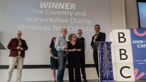 Mark Pemberton Three people collect an award on stage. There is a man standing next to them with a microphone, a woman standing behind them, and another woman standing to the side. There is a purple plinth, and a large light-up BBC sign.