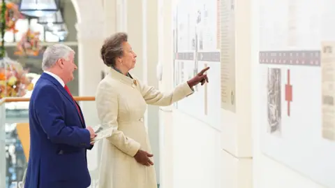 The Princess Royal viewing the Liberation Trail exhibition during a visit to St Peter Port. She is pointing at one of the boards and a man standing slightly behind her appears to be explaining it and he is holding a programme in his hand. She is wearing a long cream coat with brown gloves and he is wearing a navy suit with a red tie.