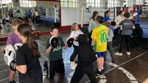 Two rows of children with boxing gloves on facing each other for sparring. There is a blue boxing ring on the right behind the children.