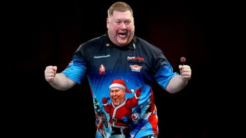 PA Media Ricky Evans celebrates with a double fist pump, with his tongue sticking out. He is wearing a bright Christmas-themed polo shirt. It appears to have a picture of himself on it, dressed as Santa holding a microphone.