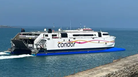 BBC Condor ferry moving through the water near a harbour. The ferry is white. Its Condor logo is printed down the side of the ferry.