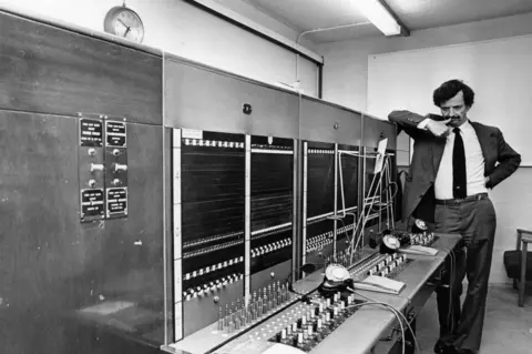 Media Wales A black-and-white photograph of a telecommunications room. A man with dark hair and a moustache stands at the far end of a large telephone switchboard that stretches across most of the image. The man is wearing a black suit. The switchboard is covered with rows of connection points, switches and patch cords, while several telephones sit on the work surface below. 