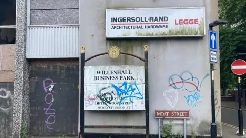 A derelict industrial building with graffiti. A sign says Willenhall Business Park, which has also been vandalised. A road sign of Moat Street is in the bottom left corner.