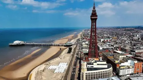 PA Media A general aerial view of Blackpool showing the tower and the town to the right, and the beach to the left with Central Pier. The sky is blue with a few white clouds.