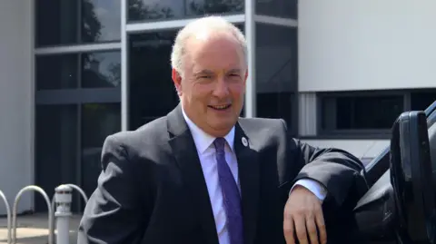 A man with short white hair leans on a van while wearing a dark-coloured suit with a purple tie.