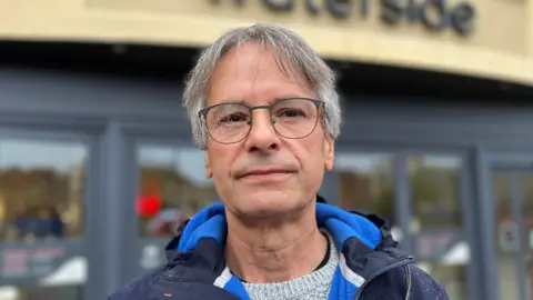 Roger Wicks. He has grey hair and grey glasses and is wearing a blue jacket. The background behind him is blurred.