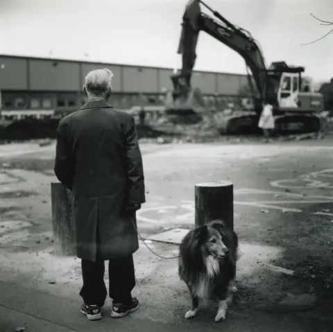 Jason Scott Tilley/Lower Block A black and white image of a man wearing an overcoat with a collie dog. He is watching as a bulldozer demolishes part of the Highfield Road site. 