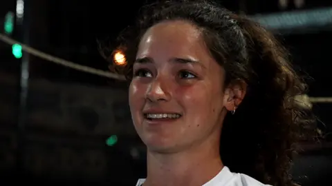John Fairhall/BBC Eira Vowles has dark hair, tied back in a ponytail, and is wearing a white T-shirt. She is smiling at the camera and is looking off to the left. 