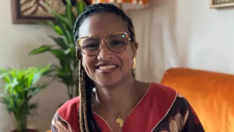 Sona Barbosa is seen wearing glasses and smiling while seated indoors, with houseplants and a sofa visible behind her.