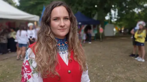 A woman with light brown curly long hair wears traditional clothes. They are an embroidered white top with a red wasitcoat.