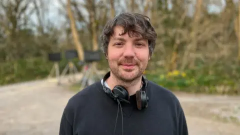 Writer Simon Mayhew-Archer on set with headphones around his neck, wearing a black jumper and trees and camera equipment in the background.
