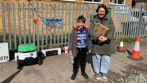 MICK LUNNEY/BBC A woman stands next to her son beside a docking station for the robots. One green and white robot can be seen and there are metal railings behind the docking station. Both mother and son are smiling widely. The woman is holding a parcel and the boy is holding a plastic water bottle.
