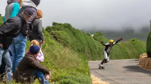 WDSC A skateboarder in black and white, and a yellow helmet, coming down the road with his hands in the air, to the left is a crowd watching.