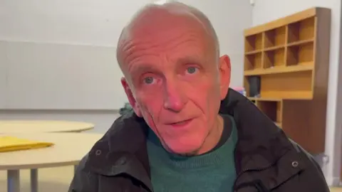Gary Spencer, who is wearing a black coat and green jumper, is sitting on a chair during a choir singing session. 