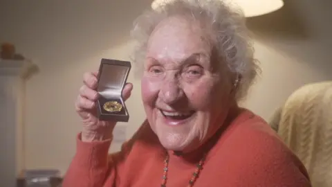 BBC Cynthia Evans looks directly at the camera, smiling. She is holding up a box containing a Bletchley Park Commemorative Badge, which is a gold medal with a blue enamel centre.