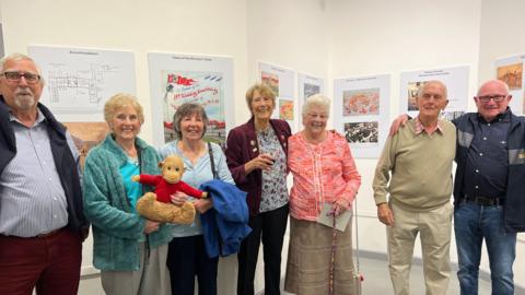 Former internees smile at the camera as they stand in front of some of the displays at the exhibition